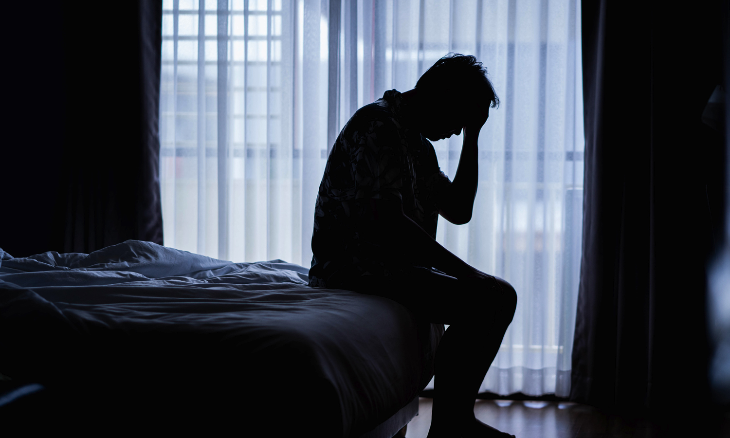 Man seated on the end of a bed in the dark.
