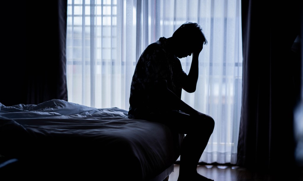 Man seated on the end of a bed in the dark.